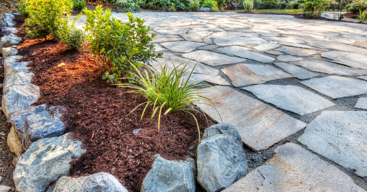 A newly-installed patio made with stone pavers with a flower bed with fresh mulch, plants, and landscaping stones.