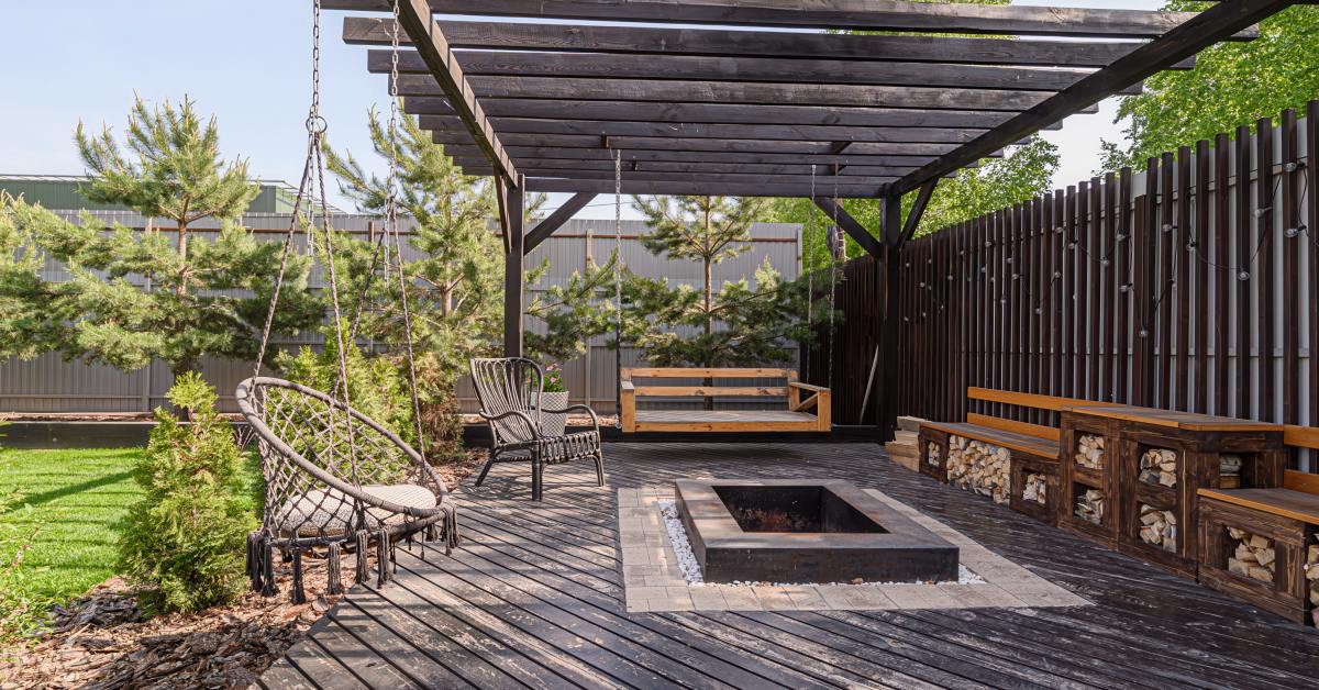An old, wooden deck area with a matching pergola overhead. There is a square, stone patio built into the center of the deck.