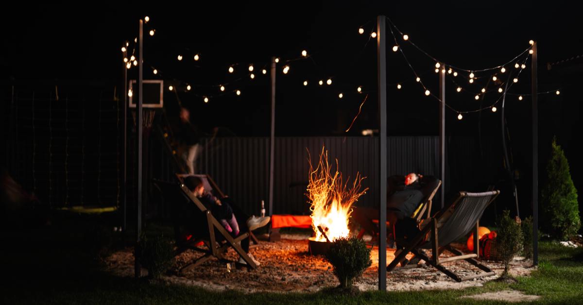 A dark night with an outdoor fire pit area in the backyard surrounded by posts holding string lights. The fire burns.