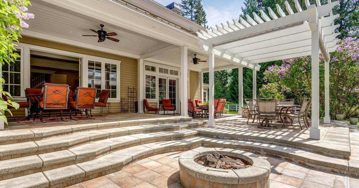A beautiful stone patio area with a white pergola and an above-ground, circular fire pit in the center of the pavers.