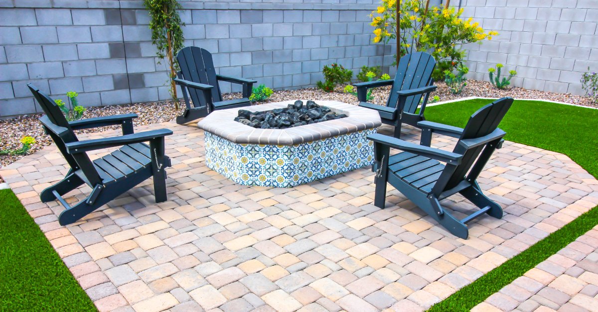 A backyard paver patio in the yard with four black Adirondack chairs and a round fire pit in the center.