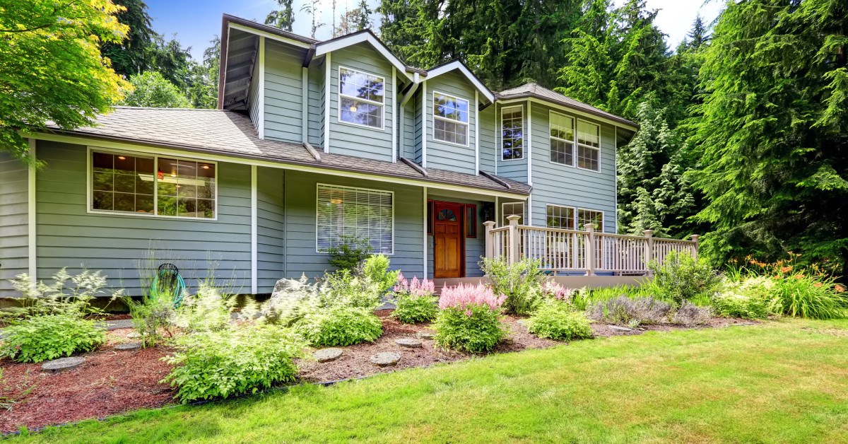 The front view of a suburban two-story home with gray paint and a wood door. The grass is cut and there are flower beds.