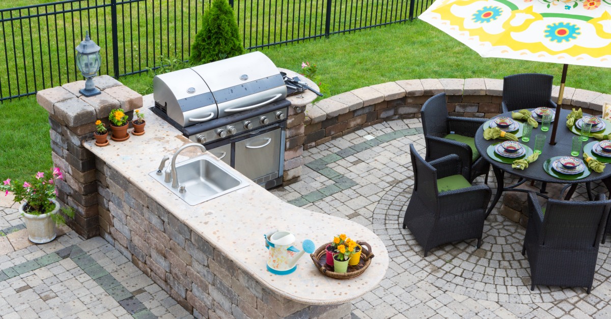 An outdoor kitchen area made of pavers, with a seating area behind the grill. There's an umbrella over the patio table.