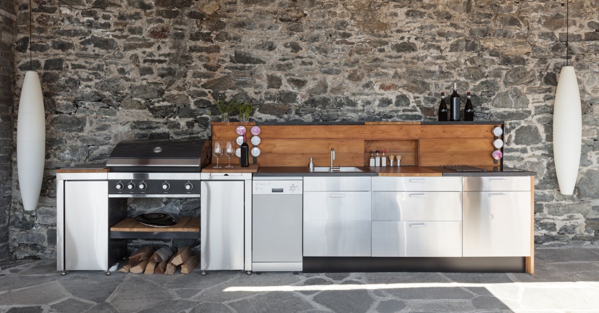 A custom outdoor kitchen with a fully stone wall and walkway. The kitchen features stainless steel cabinetry and appliances.