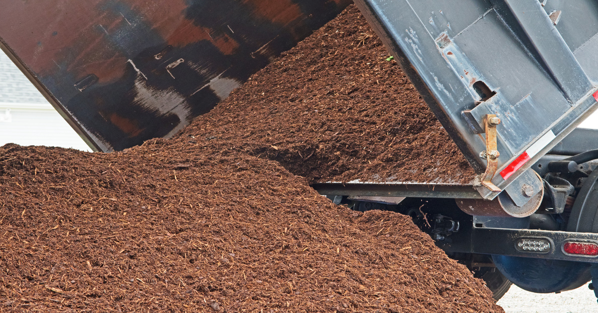 A delivery dump truck dumping a bulk landscape material delivery into a driveway. The mulch is dark red.
