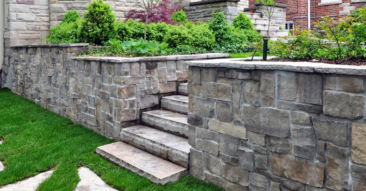 A natural stone retaining wall in a residential backyard with a set of steps in the middle, surrounding by landscaping.