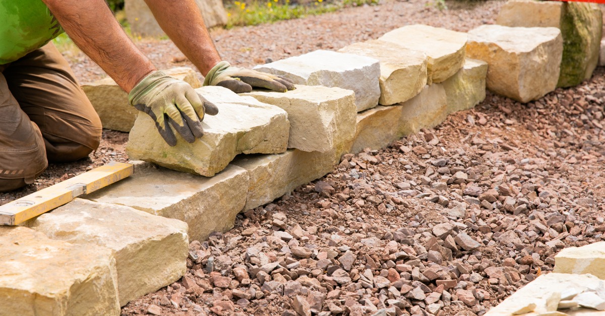 A professional landscaper on their knees, wearing garden gloves, lays landscape bricks around a new garden bed area.